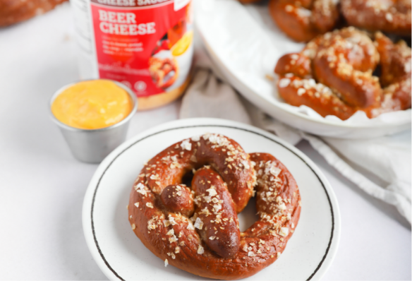 A pretzel on a white plate with a side of Hoosier Hill Farm Beer Cheese sauce on the side and a jar of powdered HHF Beer Cheese sauce