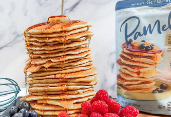 a stack of gluten free pancakes with butter and drizzled in maple syrup with a partial package of Pamela's pancake mix and some fruit in the foreground
